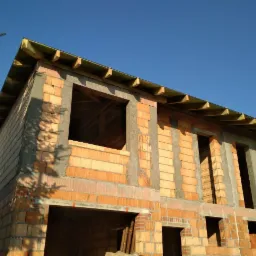 Surowy stan domu murowanego z czerwonej cegły, widoczne otwory okienne i zadaszenie, w tle błękitne niebo.