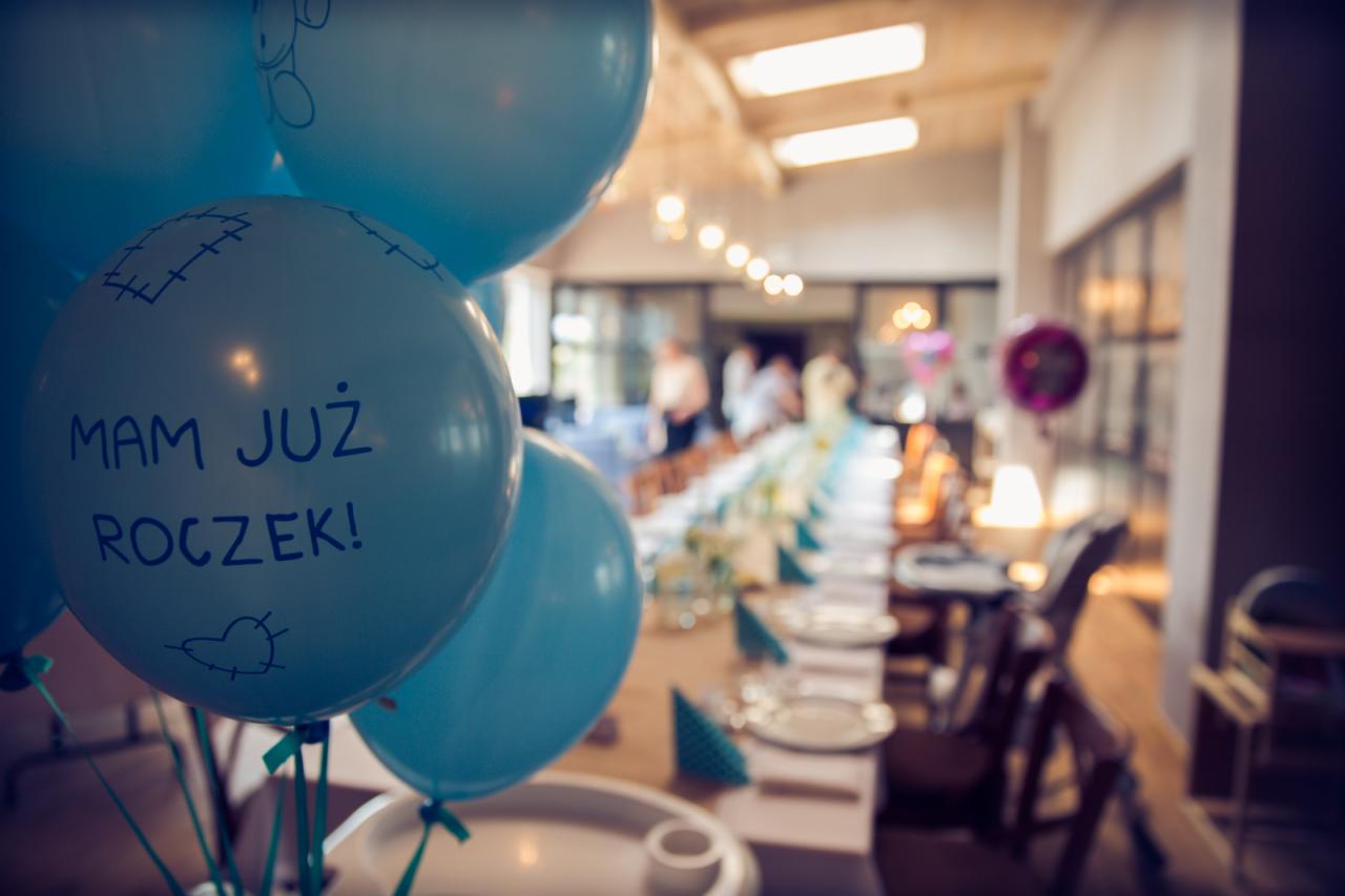 Niebieskie balony z napisem 'Mam już roczek!' na tle długiego stołu udekorowanego na przyjęcie.