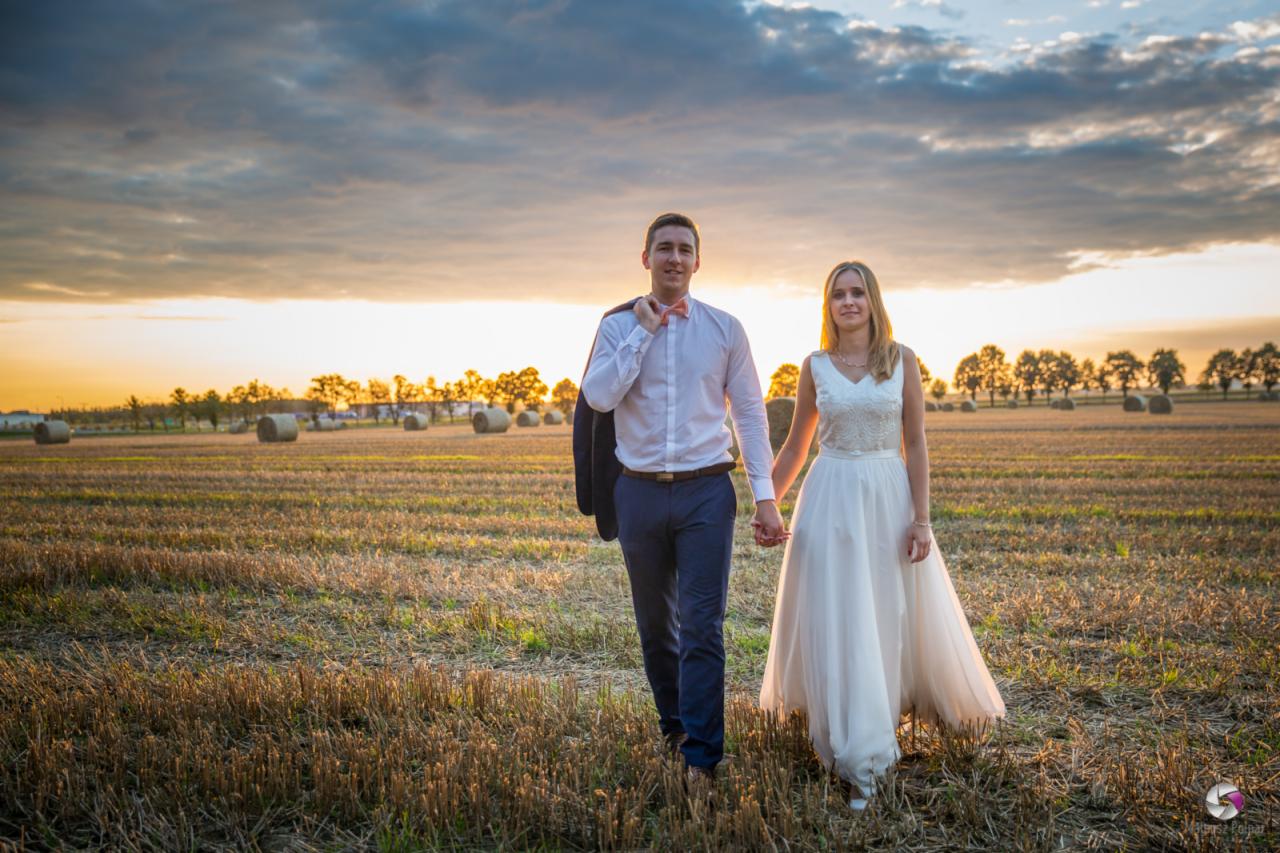 Para młoda trzymająca się za ręce spaceruje po polu ze słomianymi balotami o zachodzie słońca, mężczyzna ma marynarkę przewieszoną przez ramię.