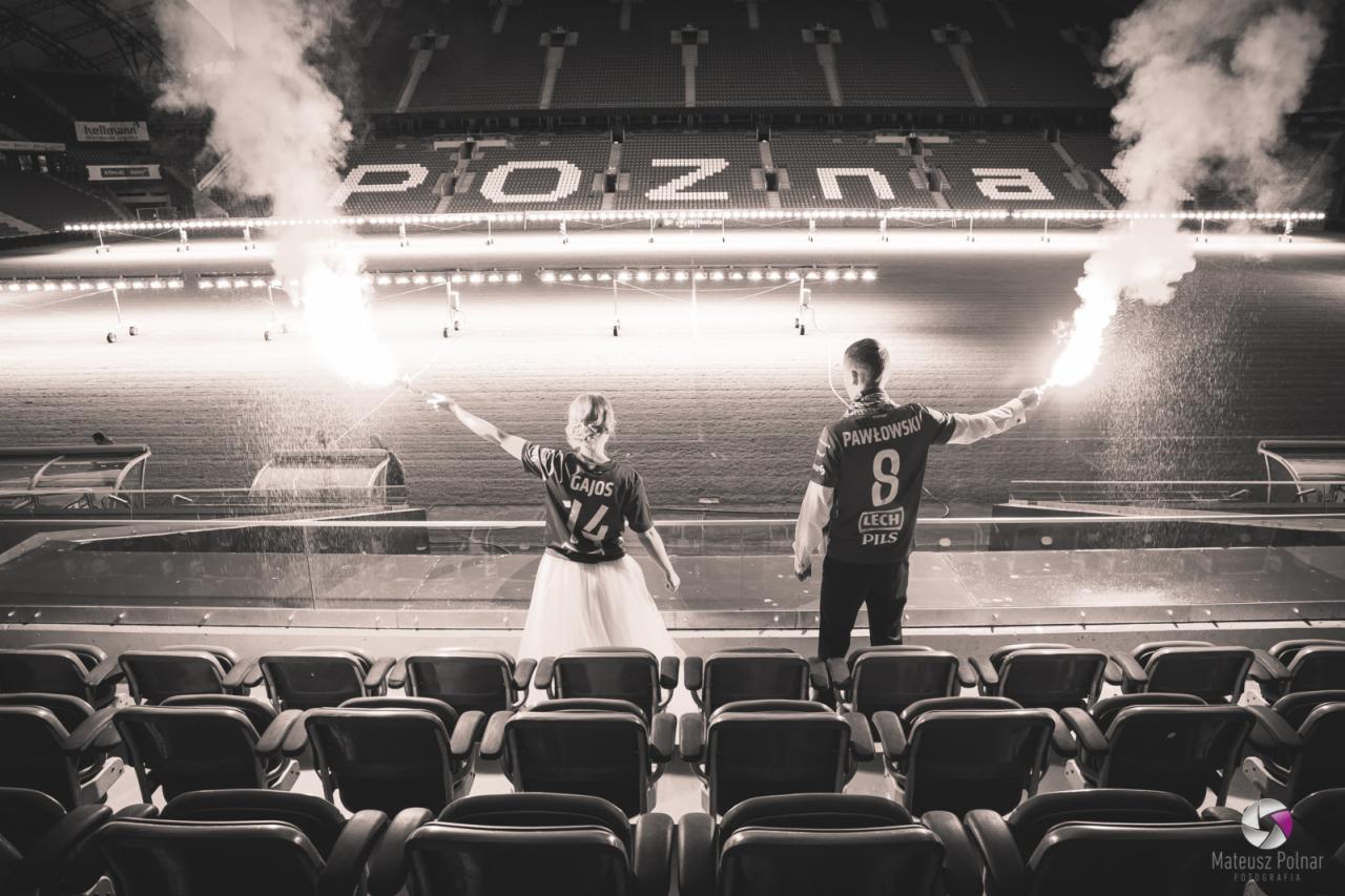 Czarno-biała fotografia pary w koszulkach Lecha Poznań z flarami na tle stadionu, dziewczyna w tiulowej spódnicy.