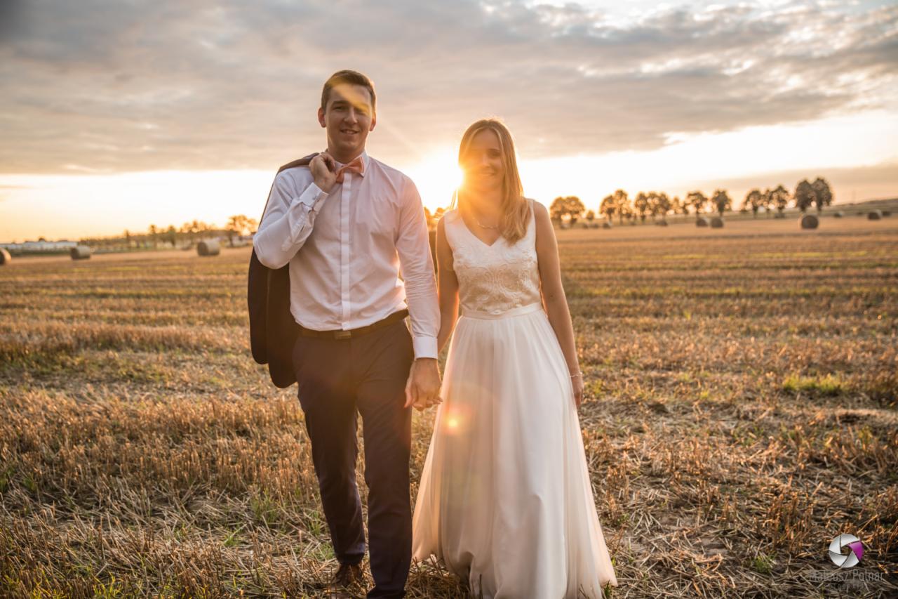 Para młoda, trzymająca się za ręce, spaceruje po polu ścierniska w promieniach zachodzącego słońca. Mężczyzna w białej koszuli z muszką i marynarką przewieszoną przez ramię, kobieta w białej sukni...