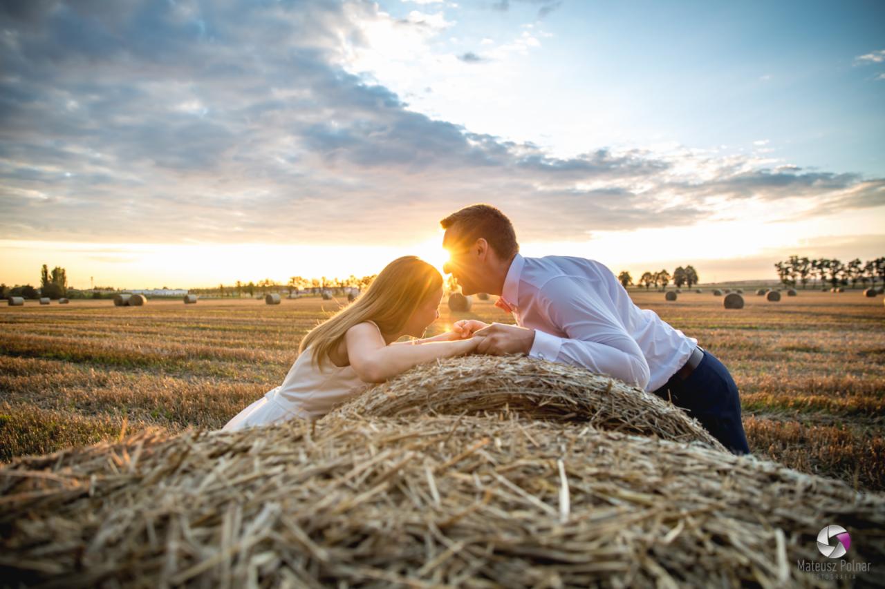 Zakochana para wpatruje się w siebie na tle zachodzącego słońca, leżąc na belach słomy na polu.