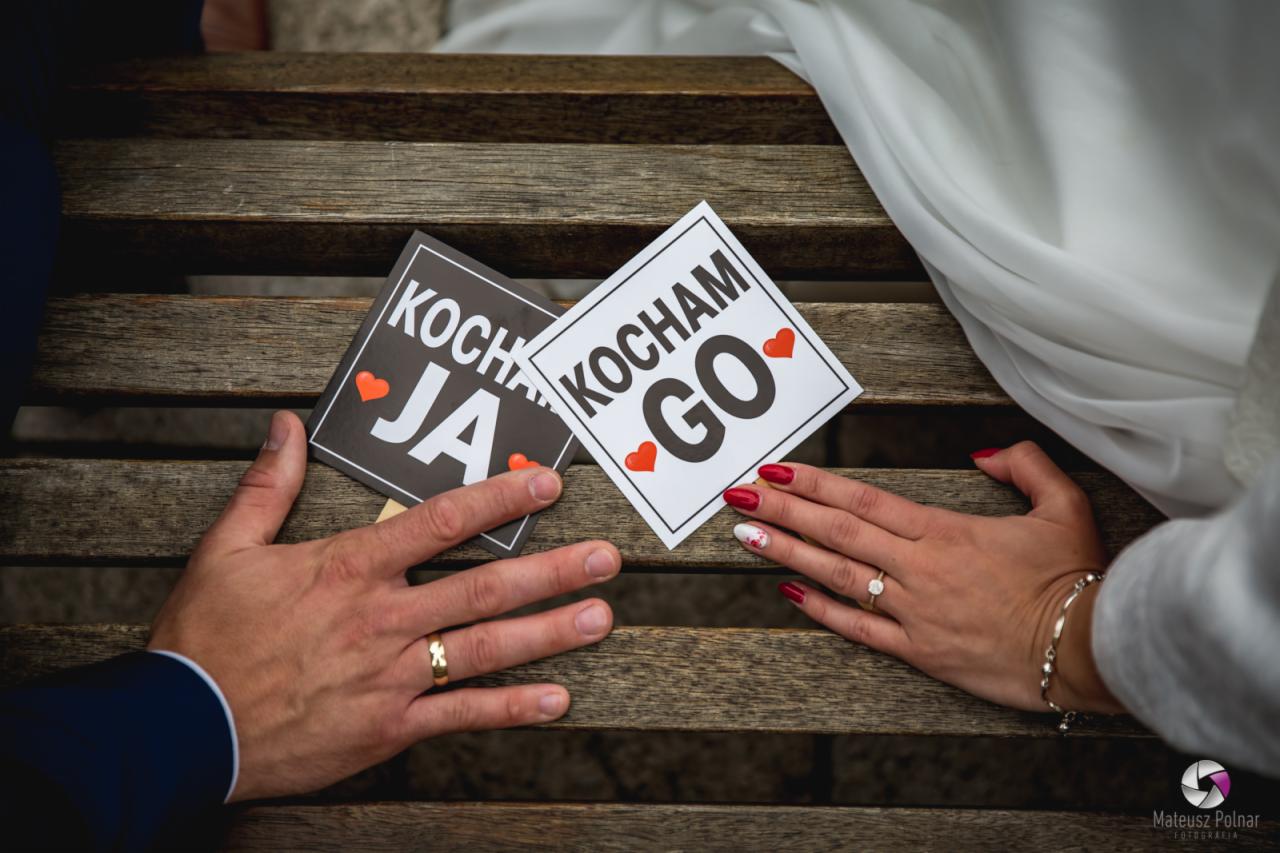 Dłonie pary młodej z obrączkami na tle drewnianej ławki, trzymające kartki z napisem 'Kocham Ja' i 'Kocham Go', uchwycone w Środzie Wielkopolskiej.