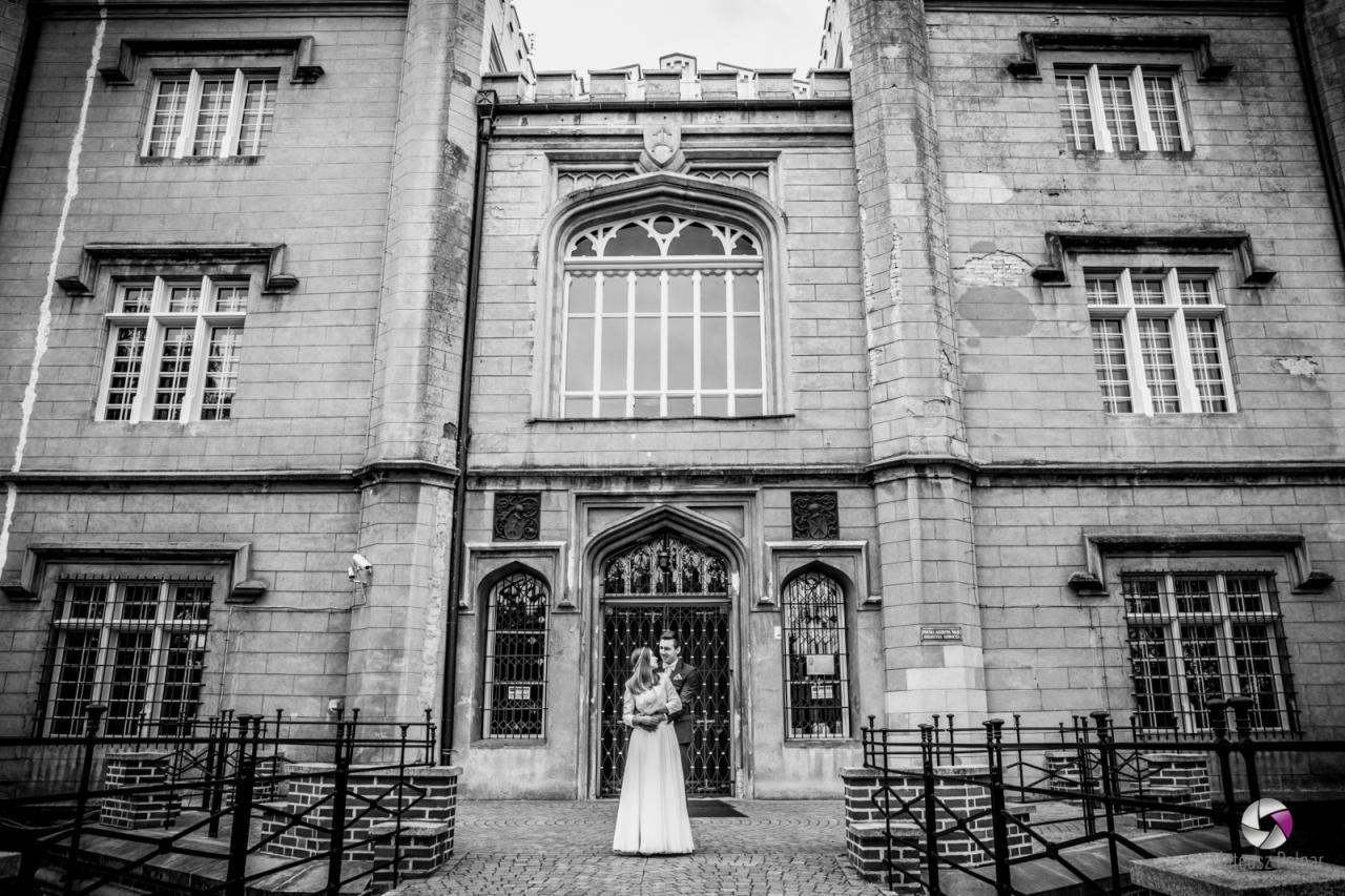 Czarno-białe zdjęcie pary młodej w objęciach przed monumentalnym budynkiem z gotyckimi oknami i dekoracyjnymi detalami architektonicznymi.
