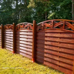 Lakierowane na brązowo drewniane ogrodzenie panelowe z ozdobnym wykończeniem górnym, widoczne z bliska na tle zieleni.