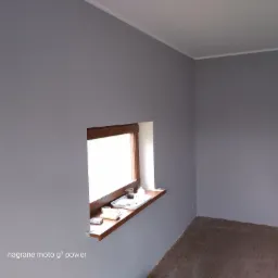 Pokój po świeżym pomalowaniu na szaro, z widocznym oknem z drewnianą ramą i parapetem, na którym leżą drobne przedmioty. Podłoga wyłożona panelami imitującymi drewno.