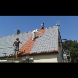 Marcin Bronowicki - Trzech mężczyzn maluje dach domu na brązowo. Jeden stoi na rusztowaniu, drugi w kombinezonie maluje dach, a trzeci stoi na szczycie dachu. Dachówka częściowo pomalowana.