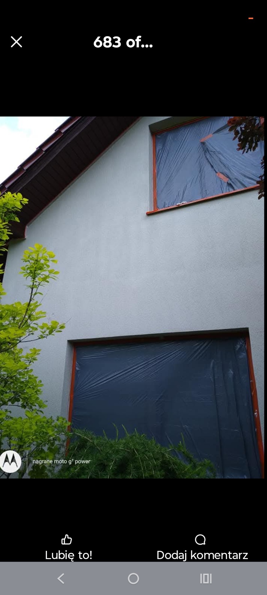Elewacja domu z oknami zabezpieczonymi folią malarską, przygotowana do prac wykończeniowych. Widoczne fragmenty zieleni. Ujęcie z dołu, lekko z lewej strony.