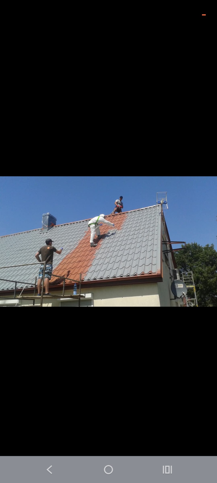 Trzech mężczyzn maluje dach domu na brązowo. Jeden stoi na rusztowaniu, drugi w kombinezonie maluje dach, a trzeci stoi na szczycie dachu. Dachówka częściowo pomalowana.