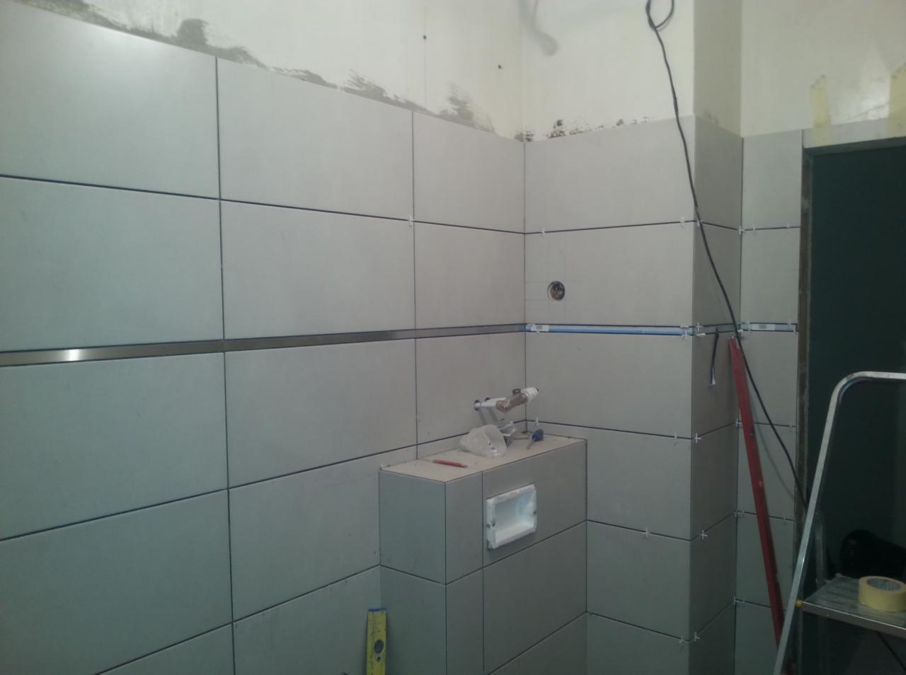 Ściana w trakcie układania jasnoszarych płytek, widoczne krzyżyki dystansowe, listwa dekoracyjna, przygotowane podejście wodno-kanalizacyjne, poziomica i drabina w tle.