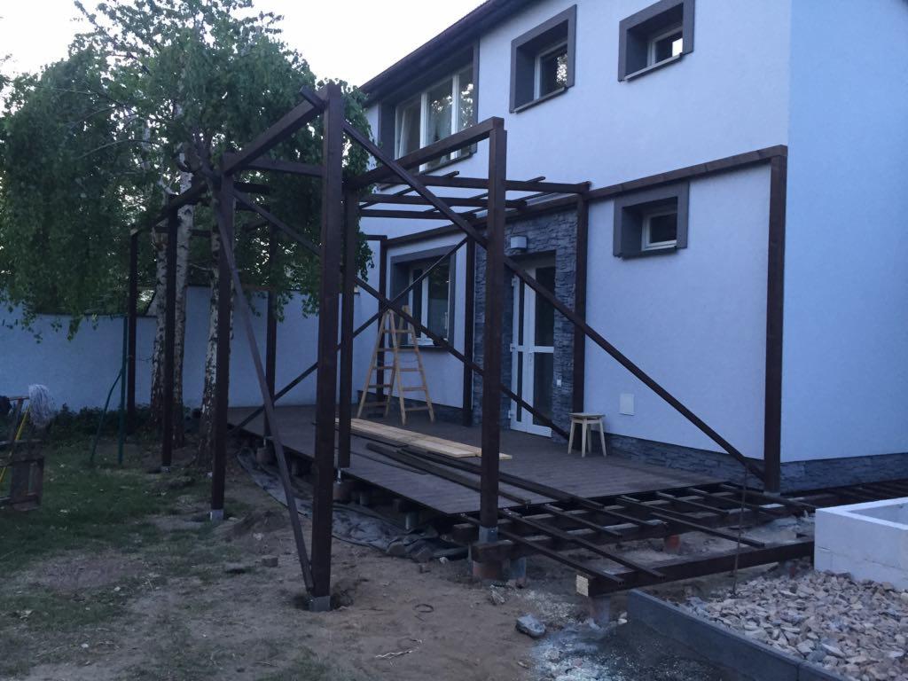 Budowa drewnianej pergoli na tarasie przy domu, widoczne elementy konstrukcyjne, deski, drabina i narzędzia, teren wokół w trakcie prac ziemnych.