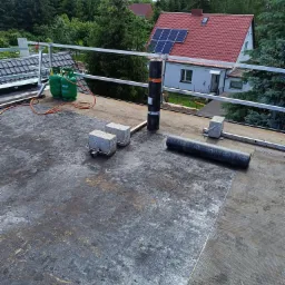 Przygotowania do krycia dachu papą termozgrzewalną, widoczne butle z gazem, rolka papy, betonowe obciążniki i częściowo oczyszczona powierzchnia dachu.