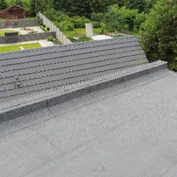 Połączenie dachu krytego szarą dachówką z płaskim dachem pokrytym papą na tle zadbanego ogrodu z altaną.