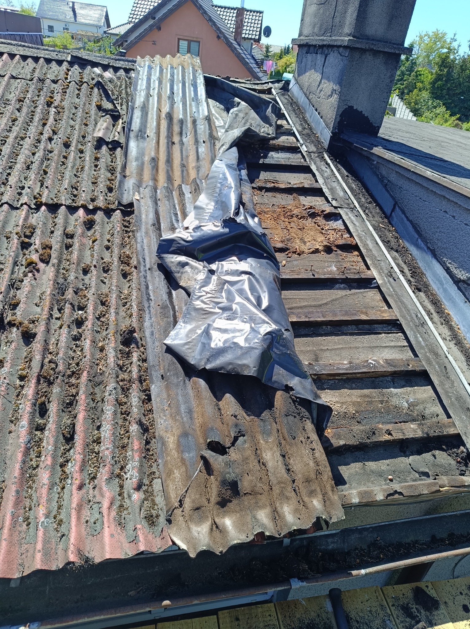 Fragment dachu z uszkodzoną, pordzewiałą blachą falistą pokrytą mchem, częściowo zakryty czarną folią ochronną, ukazujący drewnianą konstrukcję pod spodem.