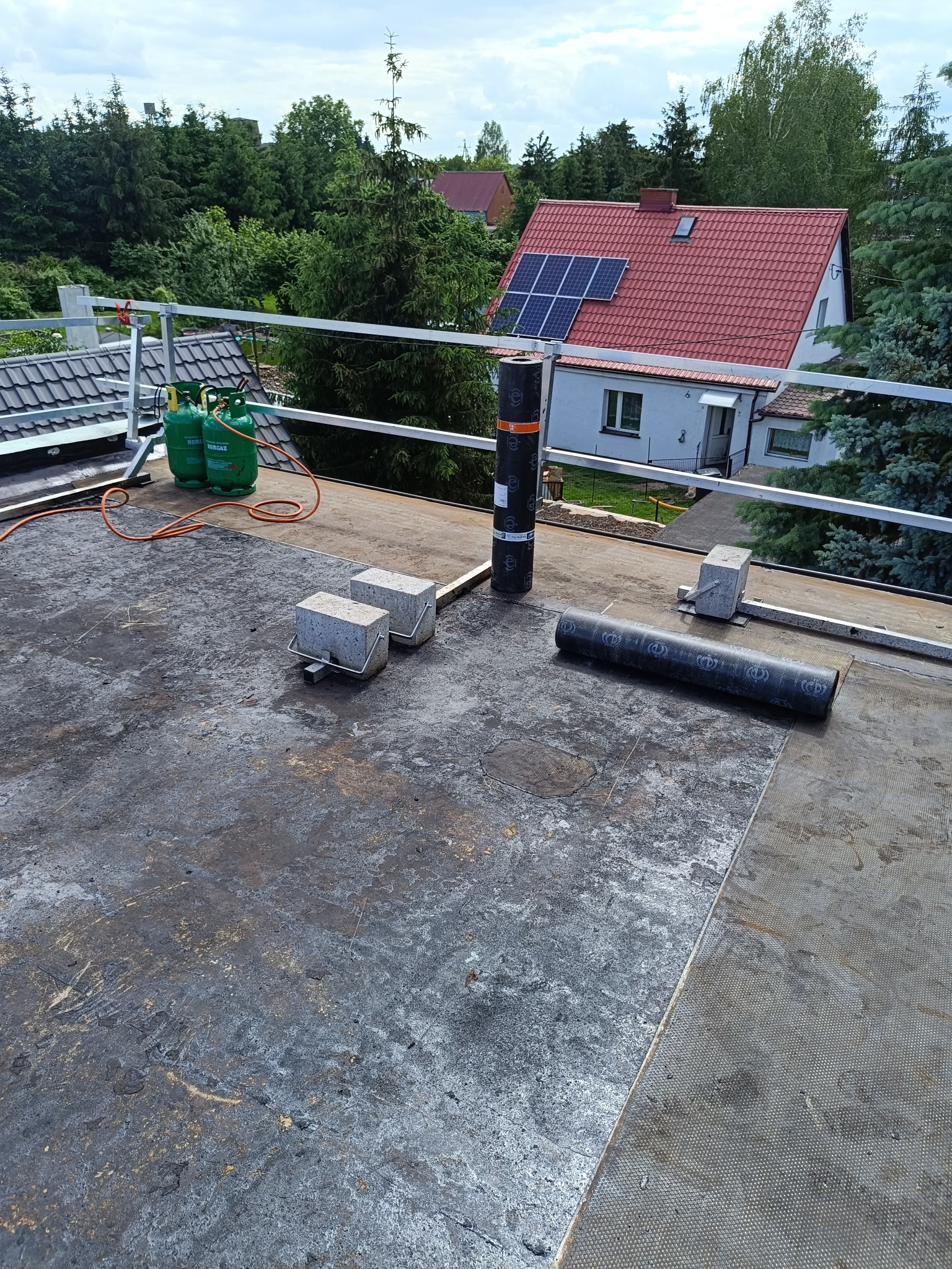 Przygotowania do krycia dachu papą termozgrzewalną, widoczne butle z gazem, rolka papy, betonowe obciążniki i częściowo oczyszczona powierzchnia dachu.