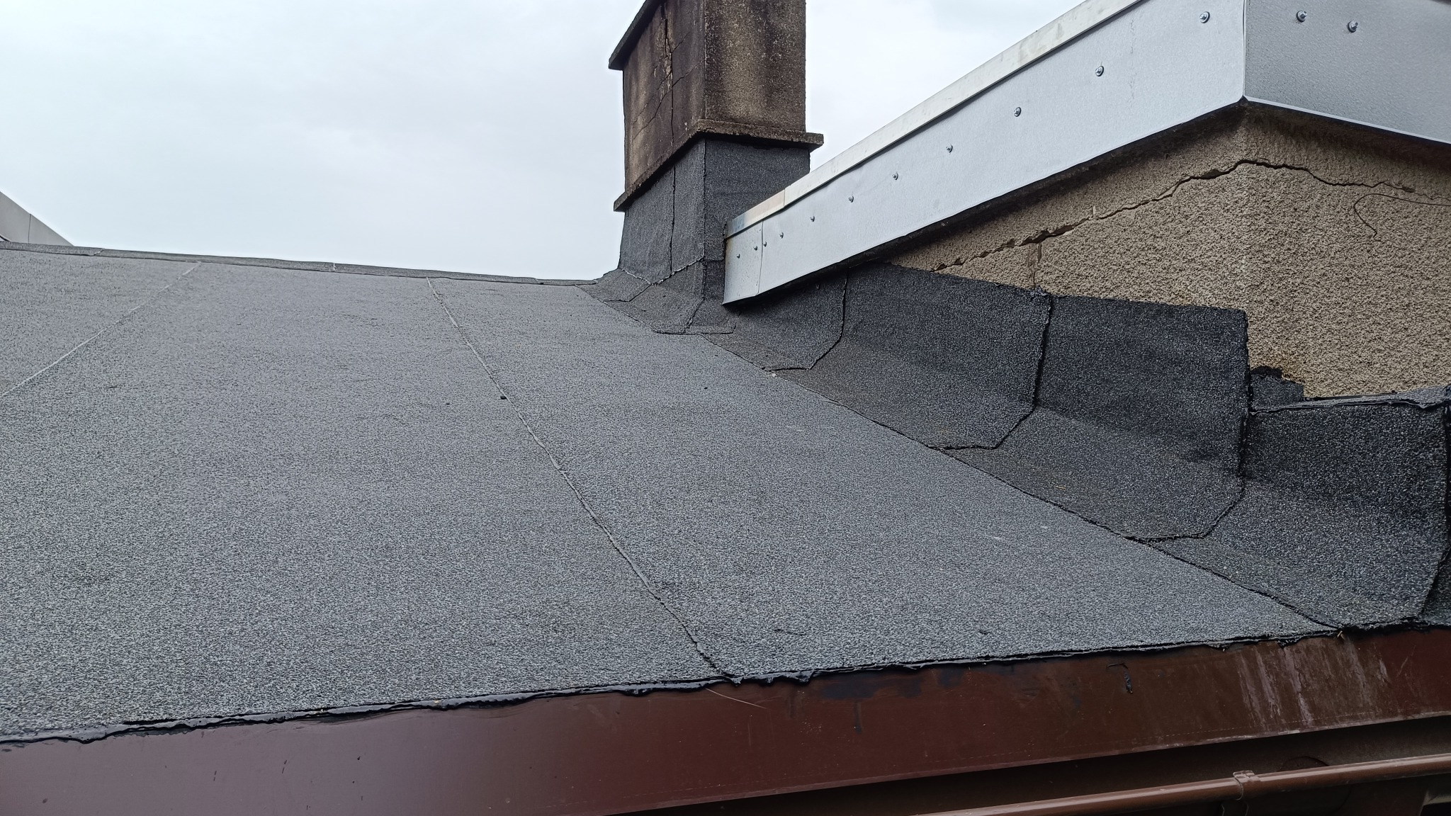 Bitumiczna papa na dachu domu z kominem i metalowym obrzeżem, widoczne pęknięcia na ścianie szczytowej.