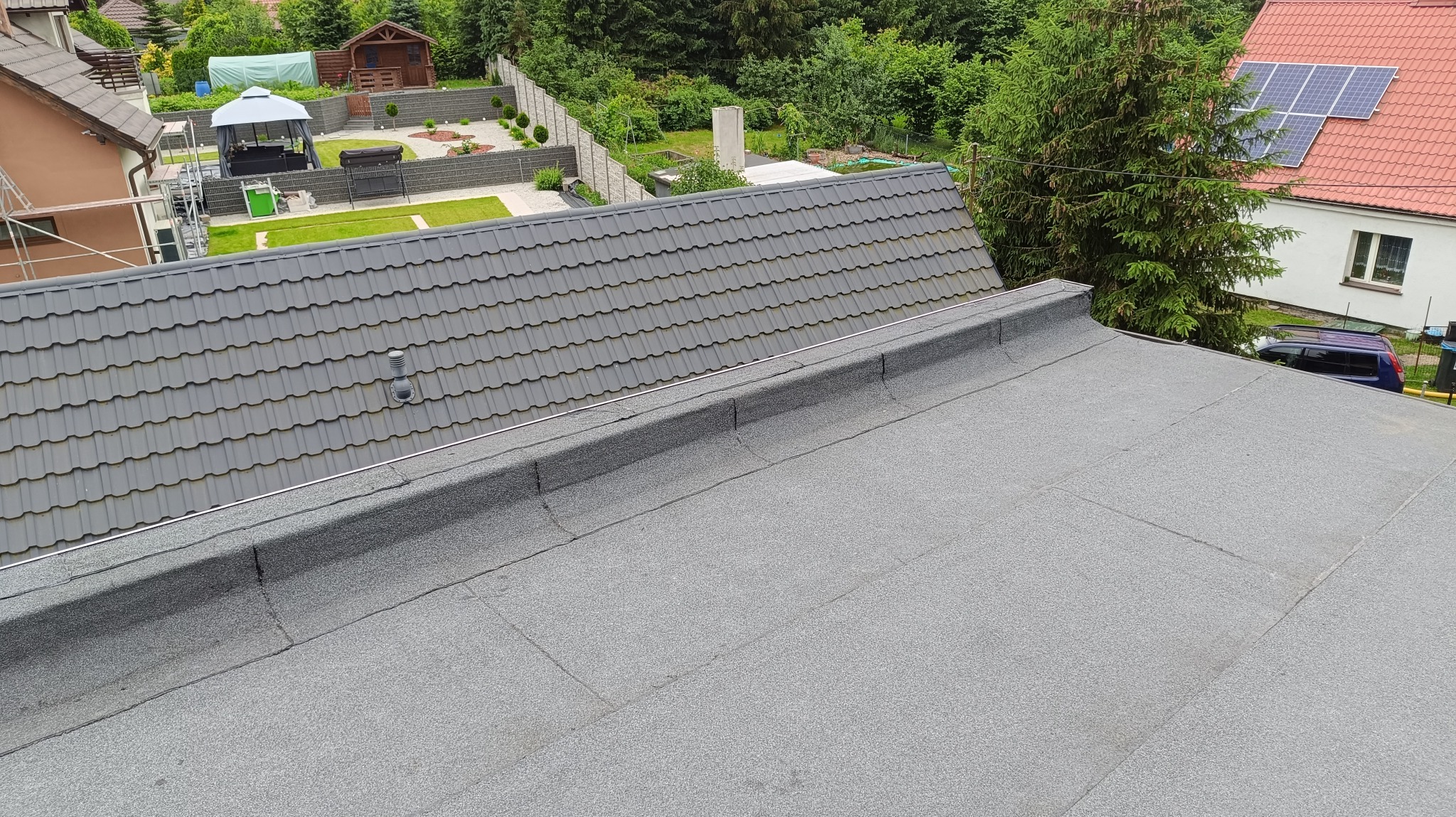 Połączenie dachu krytego szarą dachówką z płaskim dachem pokrytym papą na tle zadbanego ogrodu z altaną.