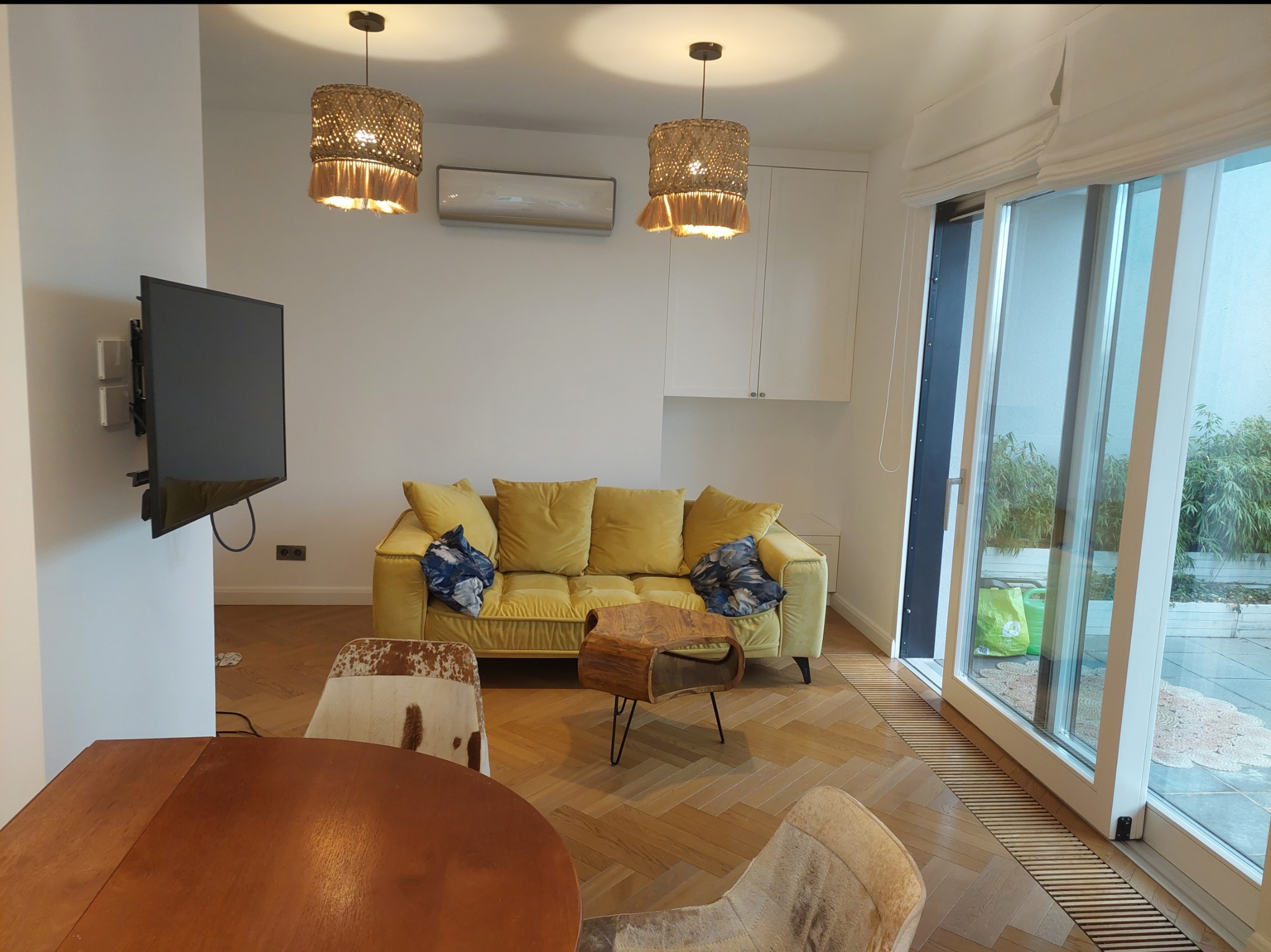 Jasny salon z żółtą kanapą, drewnianym stołem kawowym, dwoma lampami z abażurami z rafii i telewizorem na ścianie, widok na taras przez przesuwne okno.