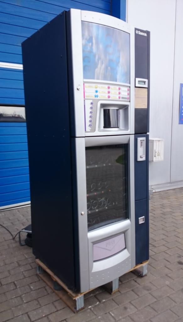 Automat do kawy i napojów Saeco w kolorze granatowo-srebrnym, ustawiony na drewnianej palecie przed niebieską bramą garażową.