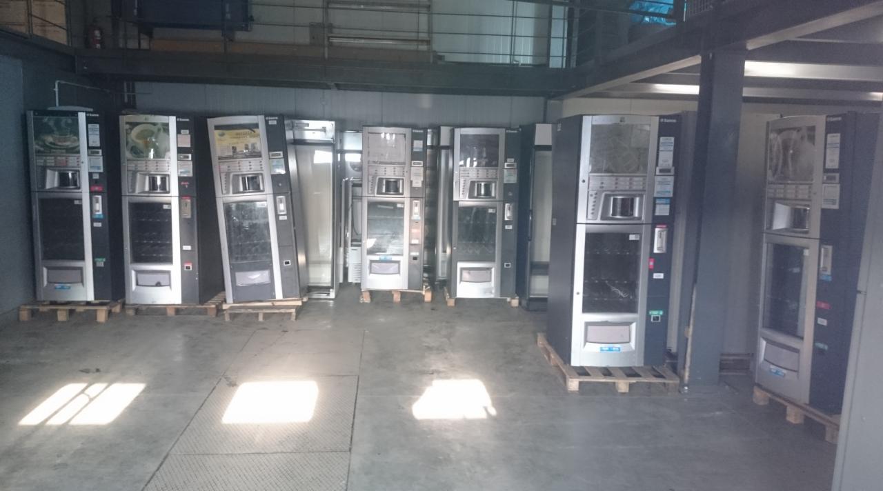 Rząd automatów z napojami i przekąskami ustawionych na paletach w magazynie lub hali produkcyjnej, widoczne metalowe elementy konstrukcyjne sufitu.
