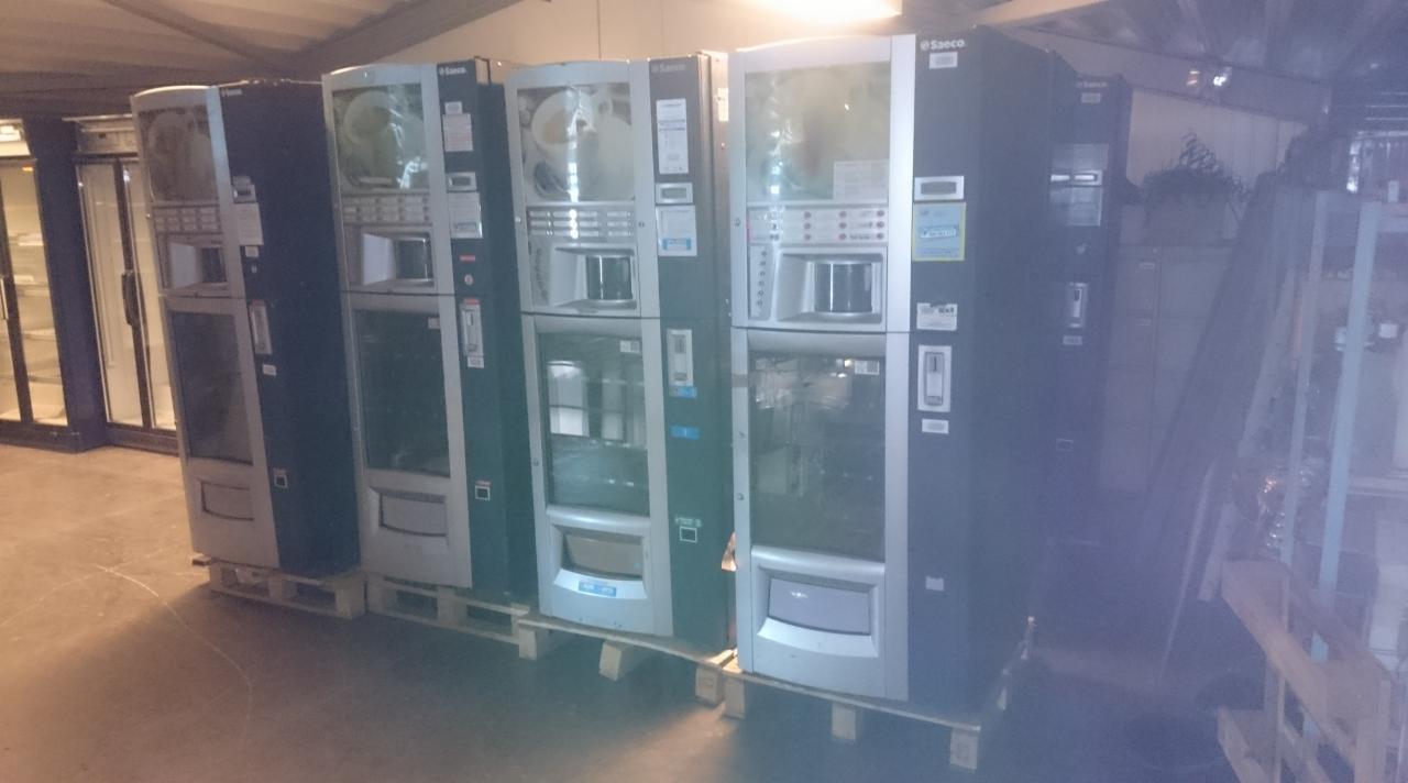 Rząd automatów do kawy Saeco, ustawionych na drewnianych paletach w magazynie, gotowych do transportu.
