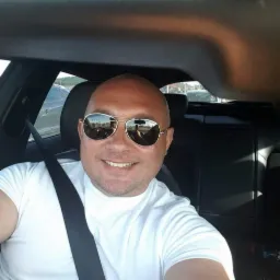 Uśmiechnięty mężczyzna w okularach przeciwsłonecznych, siedzący w samochodzie z zapiętym pasem bezpieczeństwa. Widoczne odbicia w okularach i fragment deski rozdzielczej.