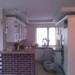 Wnętrze kuchni w trakcie prac wykończeniowych: wyspa z ceglaną okładziną, białe szafki wiszące z lampami, drabina oparta o ścianę i folia ochronna na podłodze.