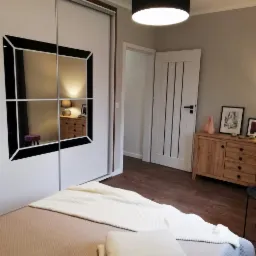 Sypialnia z łóżkiem, szafą przesuwną z lustrami z czarnymi ramami, drewnianą komodą i białymi drzwiami z czarnymi paskami.