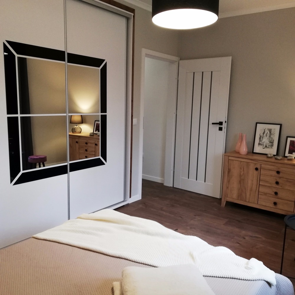 Sypialnia z łóżkiem, szafą przesuwną z lustrami z czarnymi ramami, drewnianą komodą i białymi drzwiami z czarnymi paskami.