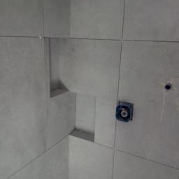 Cezary Brzozowski - Ściana wyłożona szarymi płytkami z wnęką i puszką elektryczną w trakcie montażu.