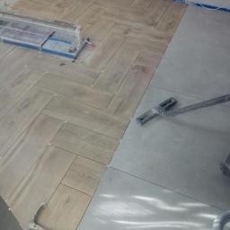 Cezary Brzozowski - Układanie podłogi: Część podłogi wyłożona jasnymi płytkami imitującymi drewno w jodełkę, druga część w szarych, gładkich płytkach. Widoczna przecinarka do płytek, młotek i odkurzacz.