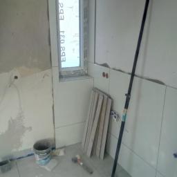 Cezary Brzozowski - Montaż płytek w łazience: widoczne białe płytki na ścianie wokół okna, podłoga z szarych płytek, wiadro po kleju, gumowy młotek i rozpórka teleskopowa.