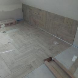Cezary Brzozowski - Układanie podłogi z płytek drewnopodobnych w jodełkę w pomieszczeniu ze skosem, widoczne kartony po płytkach i fragment ściany z ułożonymi płytkami.