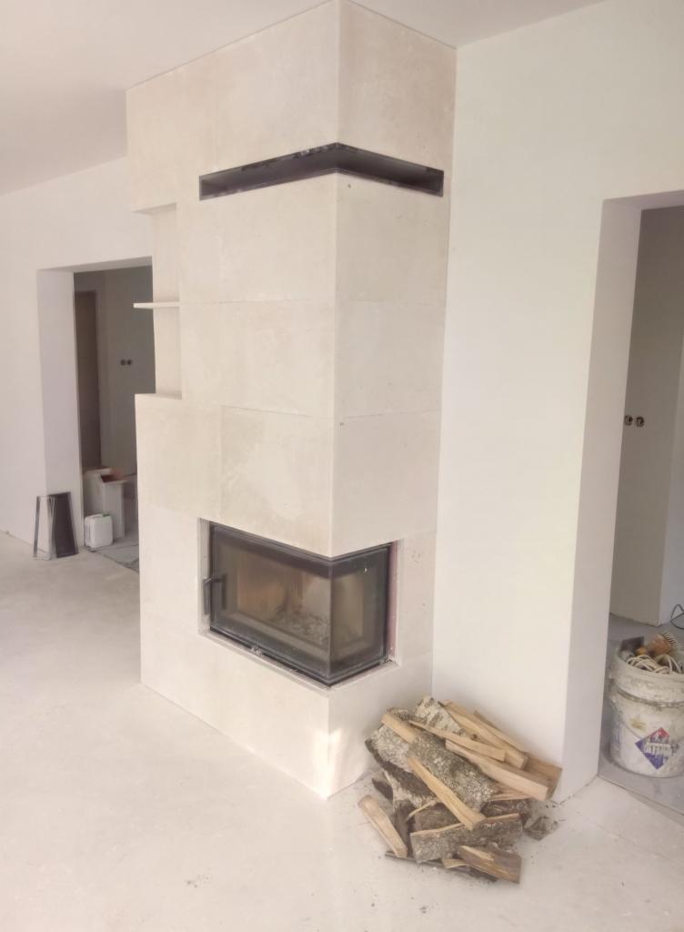 Nowoczesny kominek obłożony jasnymi płytami ceramicznymi, z widocznym paleniskiem i ułożonym drewnem obok, w surowym, minimalistycznym wnętrzu.