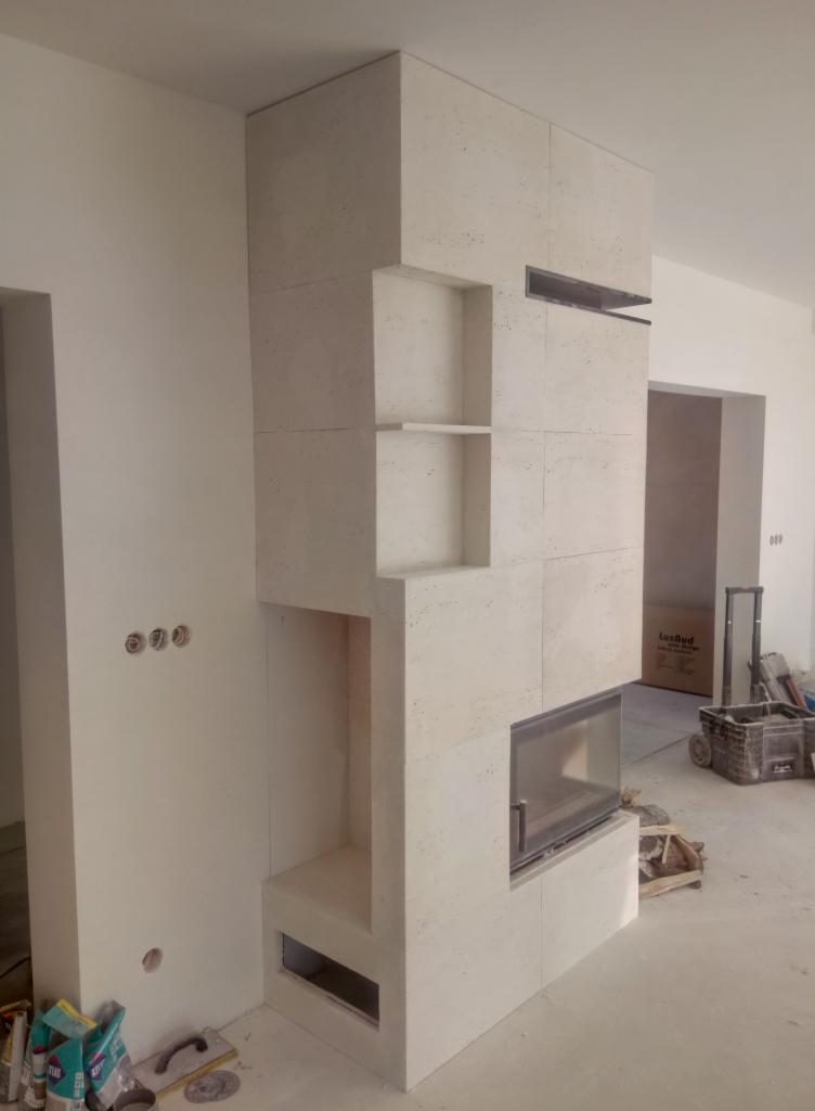 Obudowa kominka z jasnych, kwadratowych płytek trawertynowych z półkami i wkładem kominkowym, widoczne narzędzia glazurnicze w tle, surowy stan wykończenia pomieszczenia.