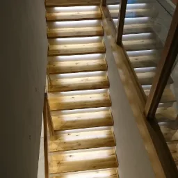 Nowoczesne schody z drewna z podświetlanymi stopniami i szklaną balustradą z drewnianymi poręczami. Ujęcie z góry, widoczne dziecięce kapcie na górnym stopniu.