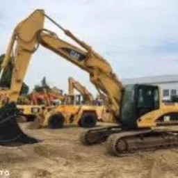 Żółty, gąsienicowy, hydrauliczny sprzęt do prac ziemnych marki CAT na placu budowy, z widocznymi innymi maszynami w tle i budynkiem.