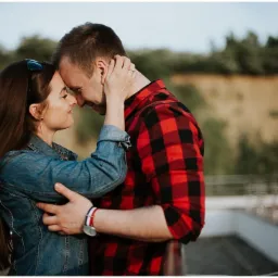 Para zakochanych z Jastrzębia-Zdroju, obejmująca się czule na tle zieleni. Kobieta w jeansowej kurtce, mężczyzna w czerwonej koszuli w kratę, dotykają się nosami.