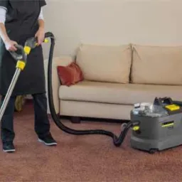 Pracownik w ciemnym uniformie czyści brązowy dywan w pokoju za pomocą odkurzacza piorącego Kärcher z żółtymi akcentami; w tle jasna sofa z poduszkami i wysokie okno z zasłonami w kwiaty.