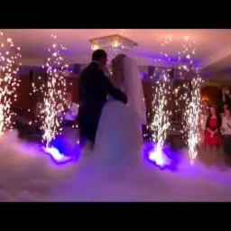 Para młoda tańczy w dymie i blasku fontann iskier podczas wesela, goście obserwują.