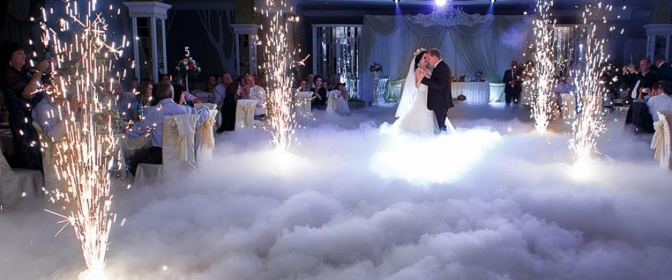 Para młoda tańczy w chmurze dymu, otoczona fontannami iskier podczas wesela w eleganckiej sali.