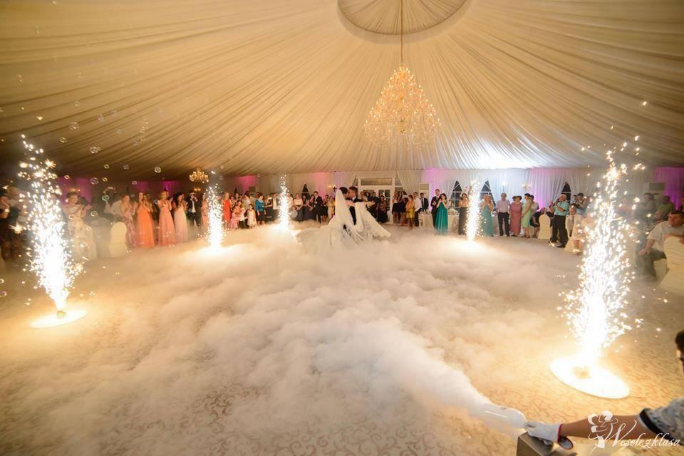 Pierwszy taniec pary młodej w dymie i blasku fontann iskier, goście w tle, sala z białym baldachimem i kryształowym żyrandolem.