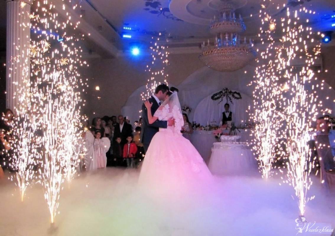Pierwszy taniec pary młodej otoczonej fontannami iskier i mgłą na sali weselnej z kryształowym żyrandolem.