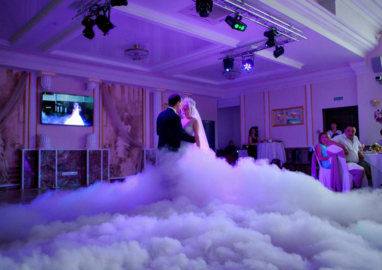 Para młoda tańczy pierwszy taniec w chmurze wytworzonej przez ciężki dym, oświetlenie sceniczne w kolorze fioletowym, goście weselni w tle.