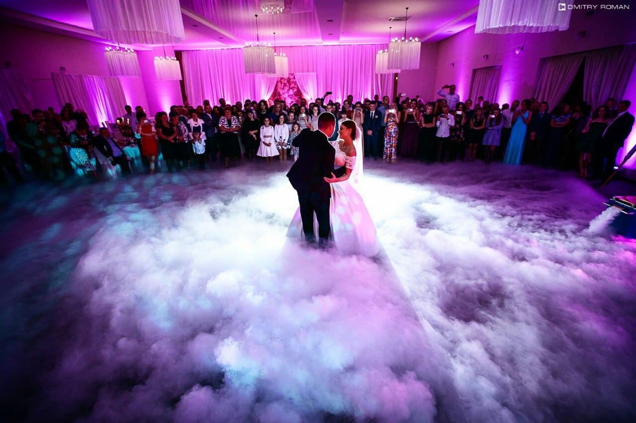 Pierwszy taniec pary młodej w sali weselnej, otoczonej gośćmi, z efektem ciężkiego dymu podświetlonego fioletowym światłem.