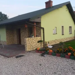 Elewacja domu jednorodzinnego z jasną, zieloną farbą i okładziną z kamienia dekoracyjnego, widoczny fragment ogrodu z dekoracjami i kwiatami w donicach.