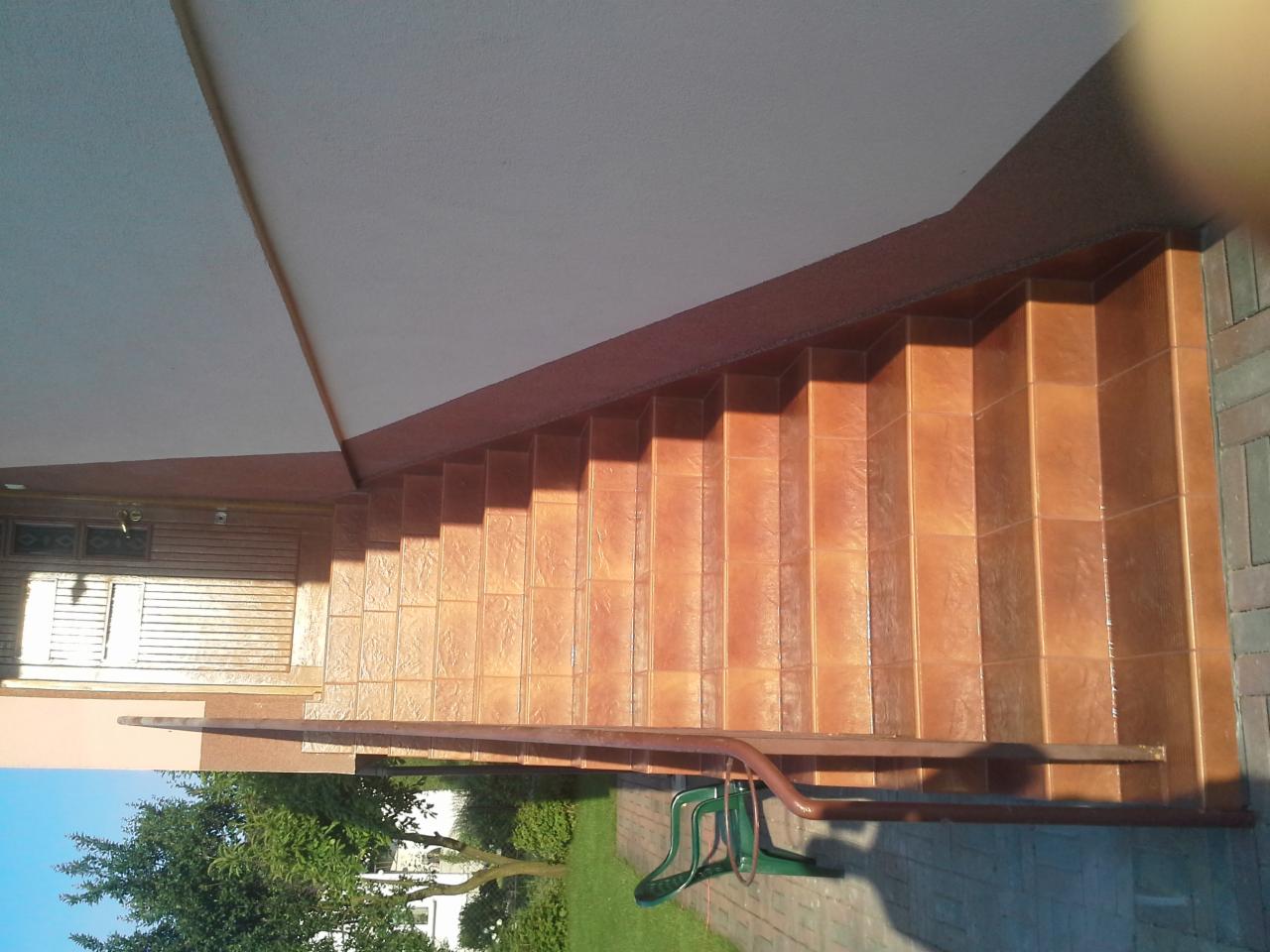 Zewnętrzne schody wyłożone błyszczącymi, brązowymi płytkami, z metalową poręczą i widokiem na zielony trawnik.