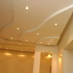 Kremowy sufit wielopoziomowy z gładkimi, falistymi liniami i punktowym oświetleniem LED, widoczne fragmenty ścian w ciepłym odcieniu beżu.
