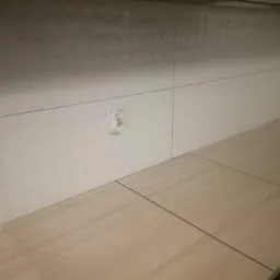 Białe, wzorzyste płytki ścienne w kuchni z gniazdkiem elektrycznym, jasny blat kuchenny z widocznymi fugami i ciemne szafki wiszące.