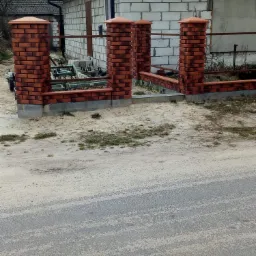 Ceglany płot z czterema słupkami i metalowymi poprzeczkami przed budynkiem z białej cegły, widoczne maszyny rolnicze w tle.